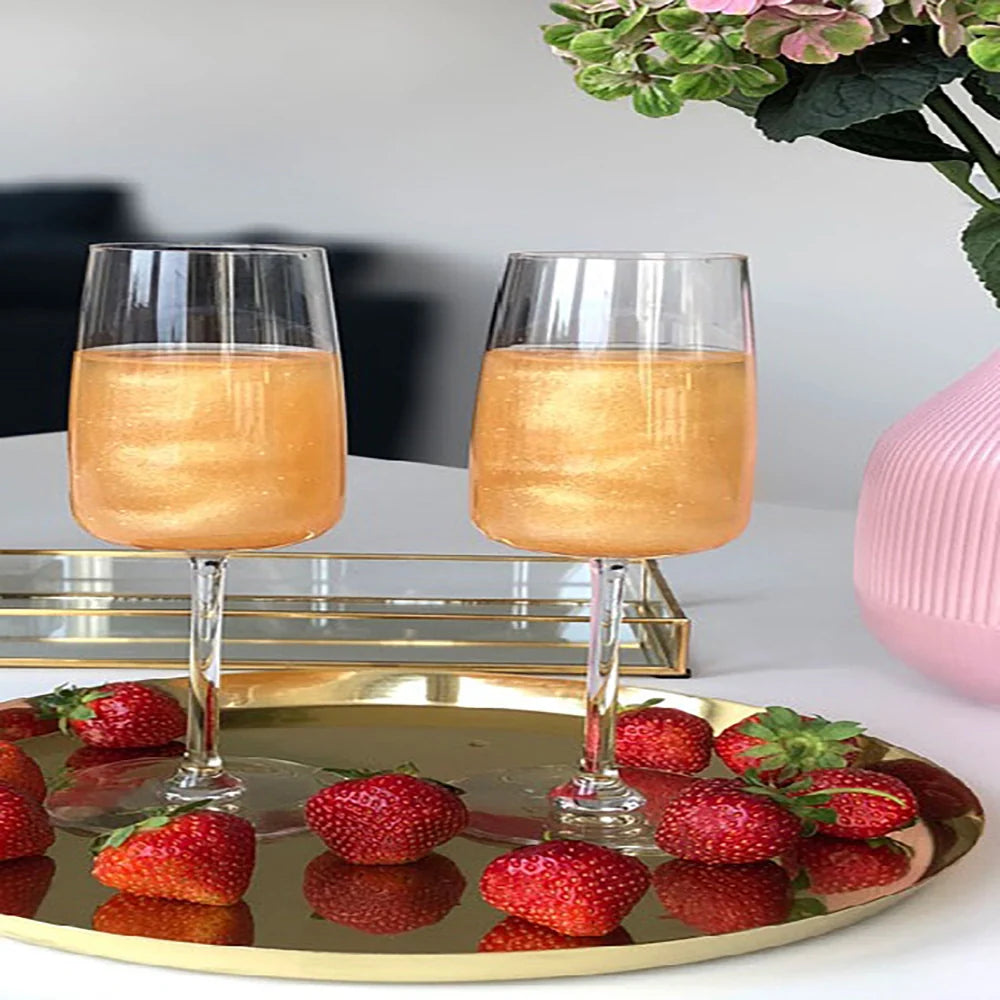 Ätbara massageoljor med jordgubbsmak – två glas med guldfärgad olja omgivna av färska jordgubbar på ett gyllene fat, tillsammans med rosa blomster i bakgrunden från Chambers of Love