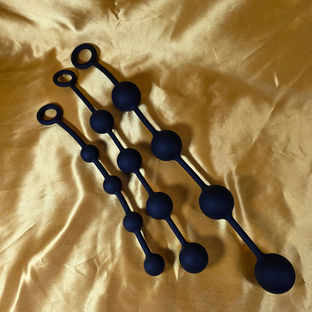 Fyra Kulors Anal Beads i tre storlekar – liten, mellan och stor – i silikon med stegrande kulor och säkerhetsring, fotograferade på guldsidenbakgrund. Från Chambers of Love.